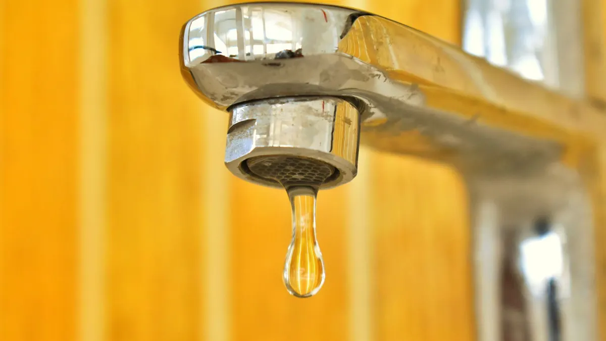 A close-up shot of a water droplet hanging from a stainless steel faucet, highlighting surface tension.