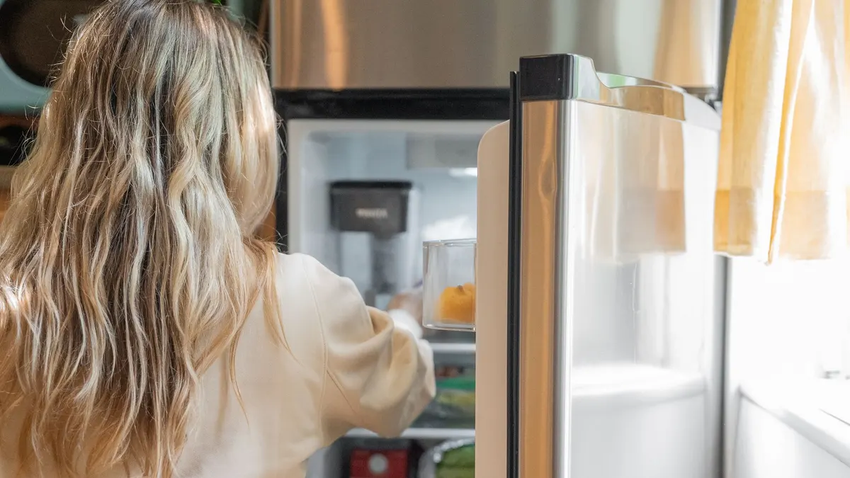 Back view of a blonde woman retrieving food from a refrigerator in a bright kitchen.