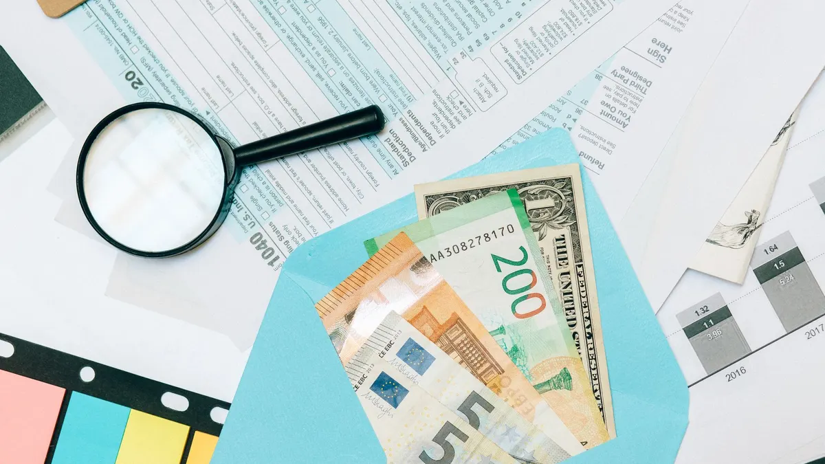 Close-up of tax documents and mixed currency in a blue envelope with a magnifying glass, perfect for financial analysis concepts.