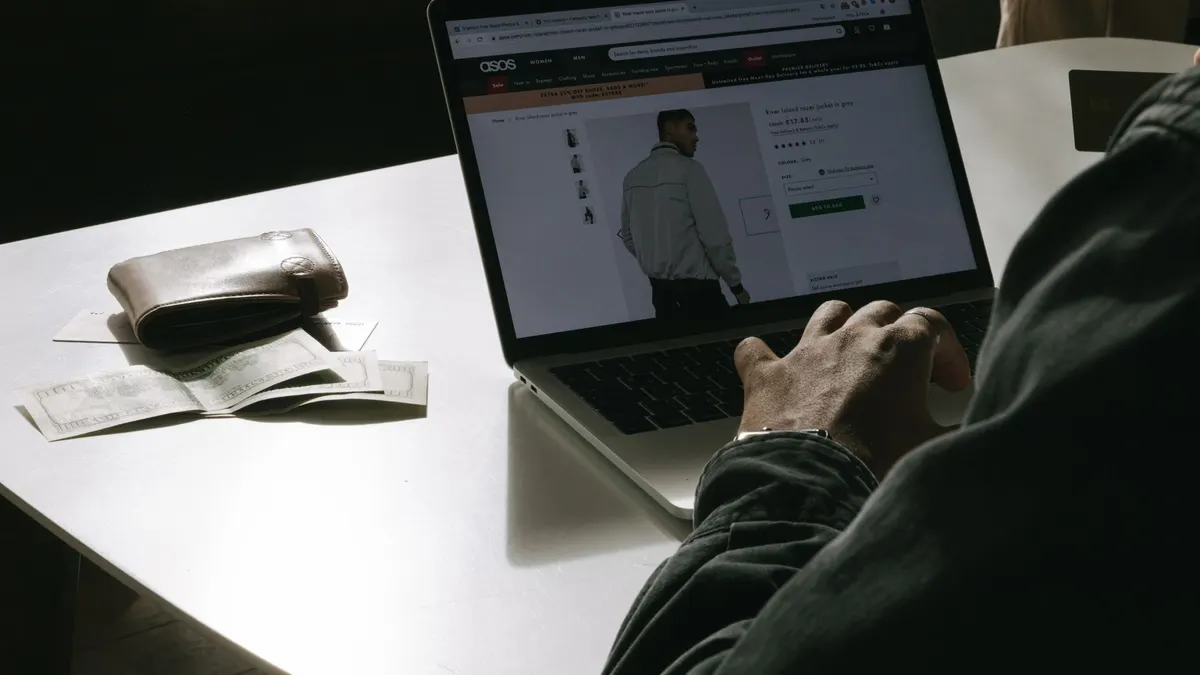 Adult man shopping for clothes online at home using a laptop, with a wallet on the table.