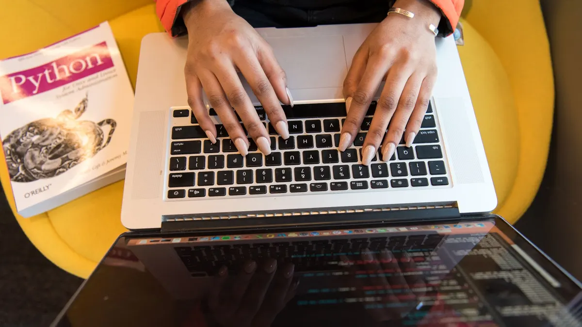 Close-up of hands typing on a laptop keyboard, Python book in sight, coding in progress.