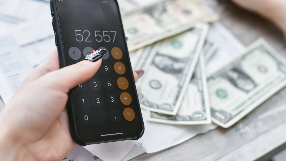 Close-up of hands using smartphone calculator next to dollar bills on table. Financial planning concept.