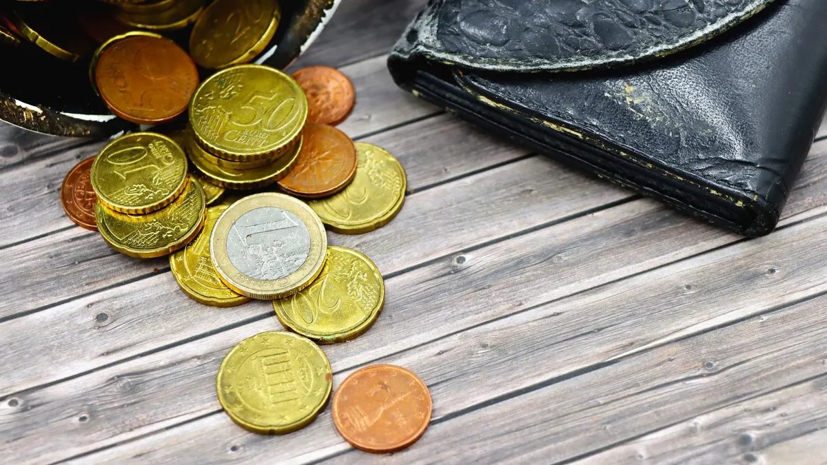 A worn-out leather wallet and scattered euro coins on wooden surface suggest economic hardship.