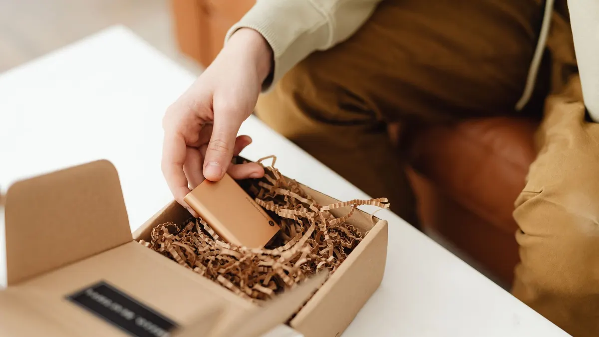 A person unboxes a small item from a cardboard package filled with crinkle paper, indoors.