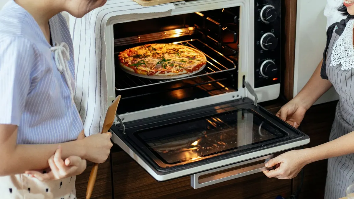 Side view of anonymous female cooks in aprons placing delicious homemade pizza in hot electrical oven while baking in kitchen