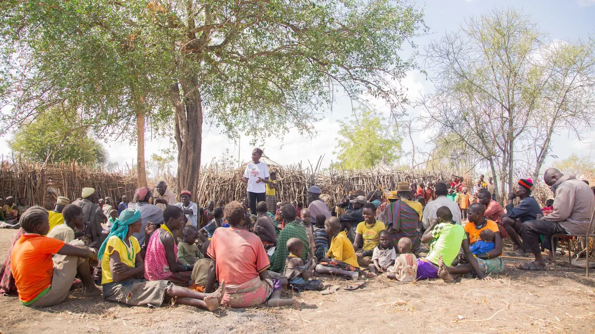 A diverse group of adults and children gather under a tree for a community meeting outdoors.