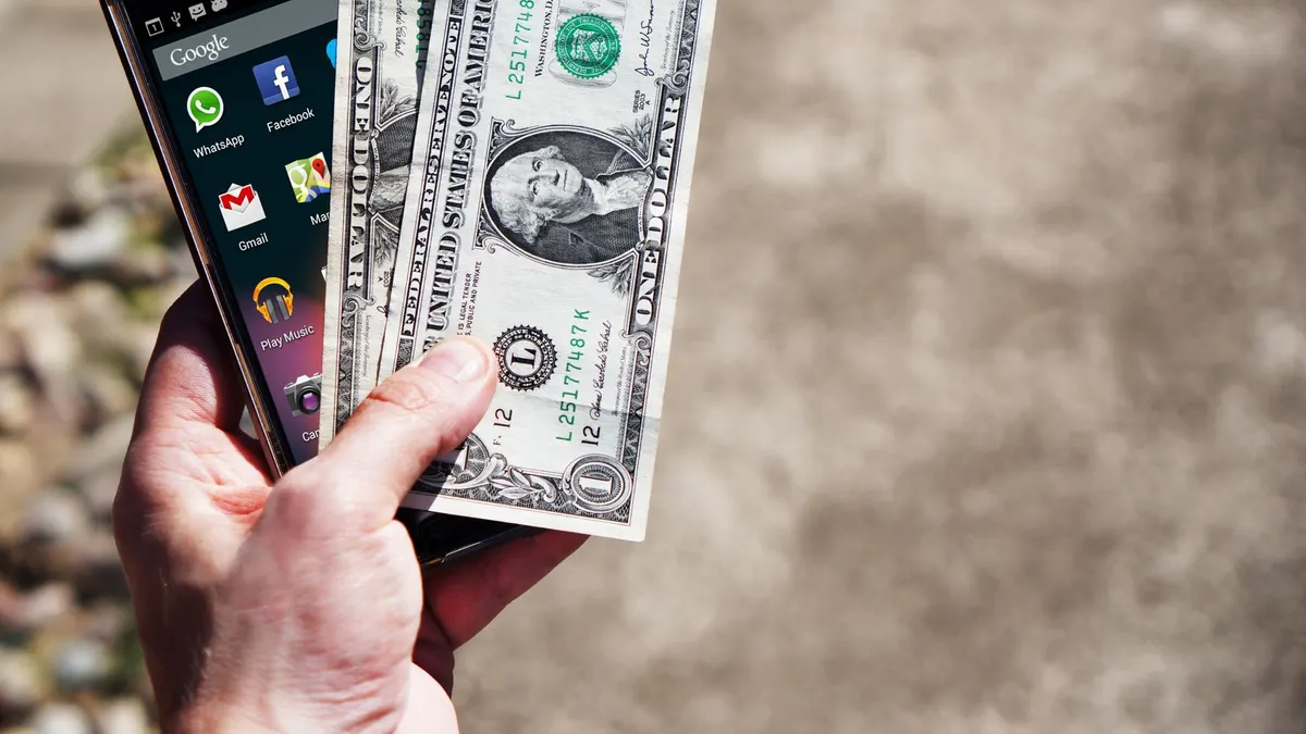 Close-up of a hand holding US dollar bills and a smartphone outdoors, showcasing financial technology.