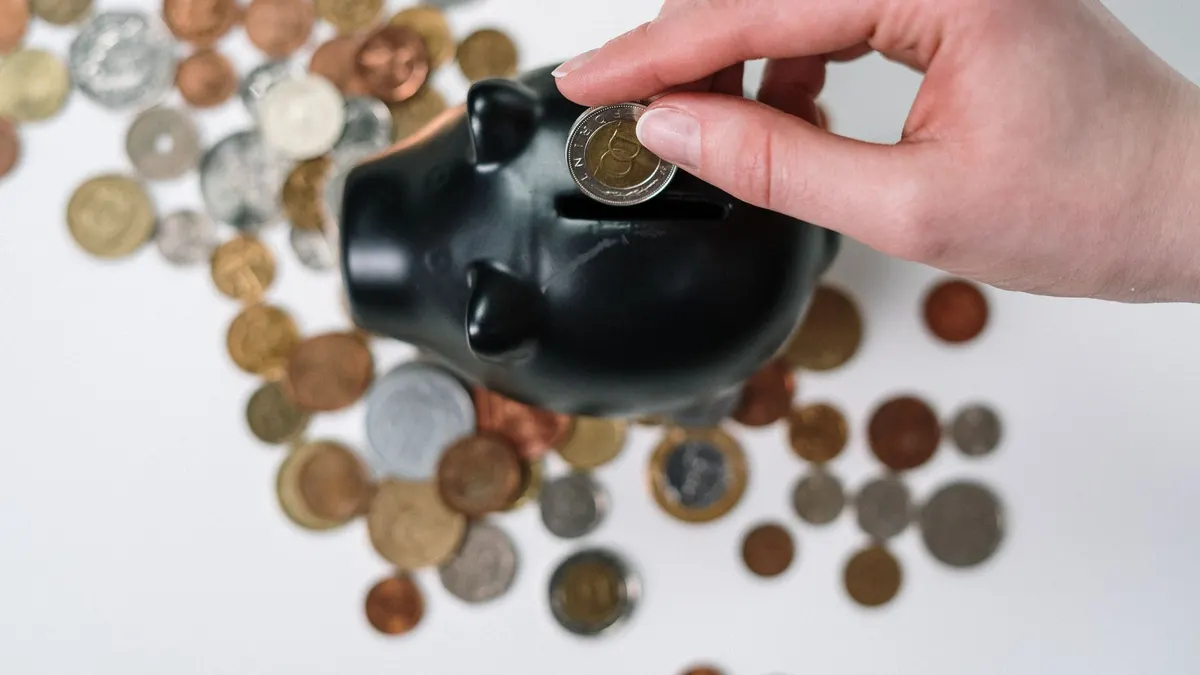 Close-up of a hand inserting a coin into a black piggy bank with scattered coins on a white background.