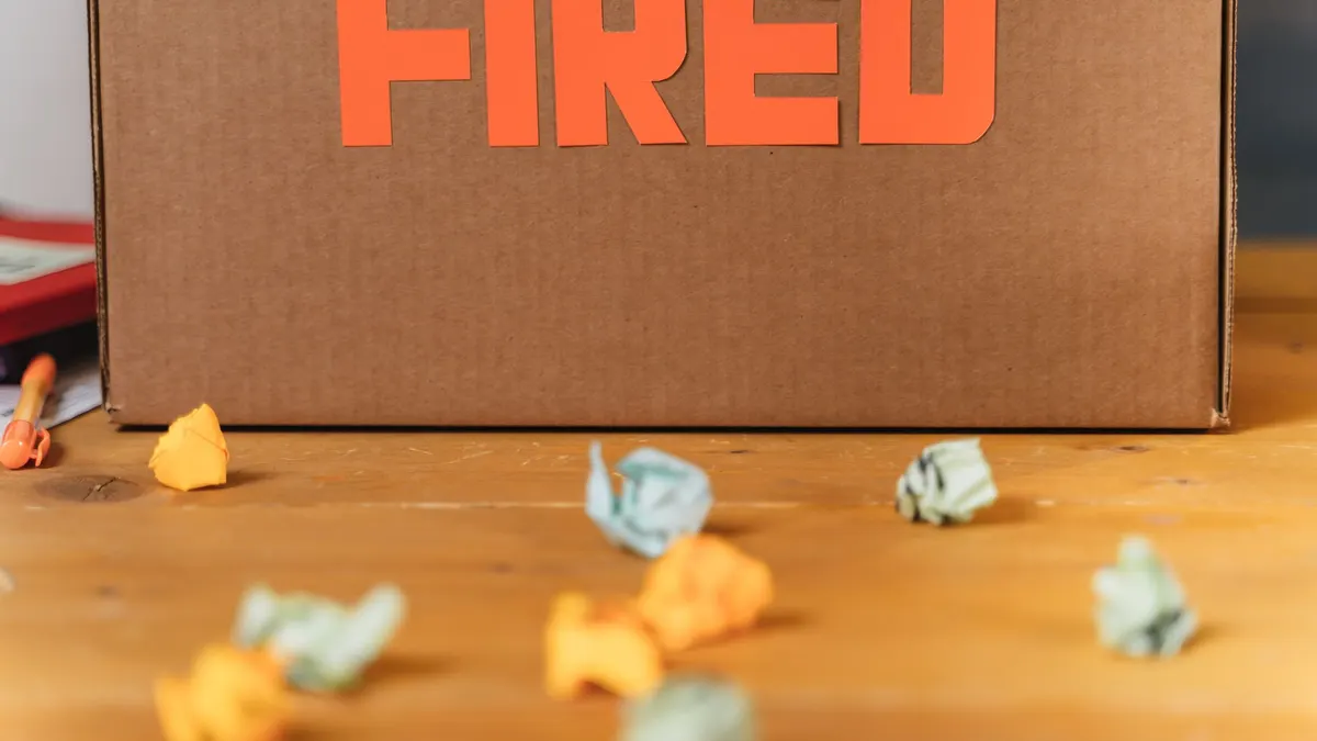 Close-up of a cardboard box labeled FIRED on a wooden table with crumpled papers.