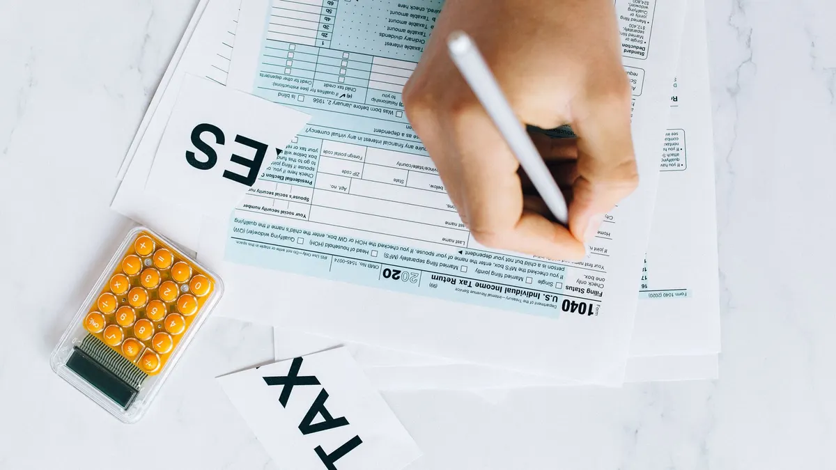 Close-up of hand writing on tax form with calculator nearby on white surface.