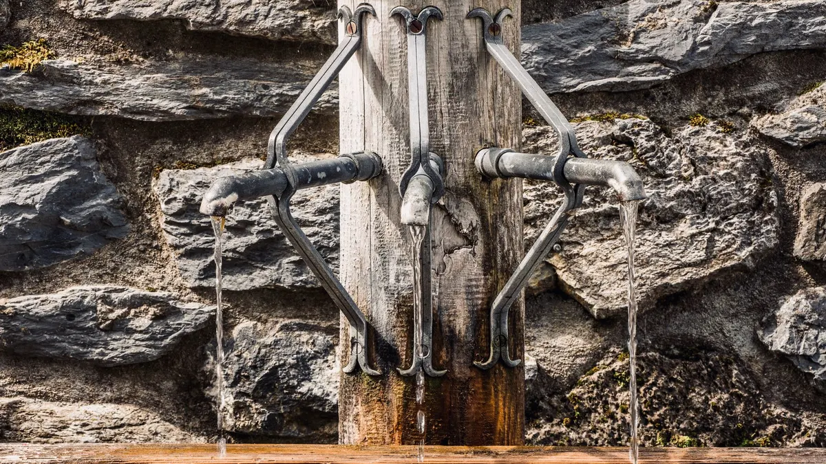 Metal taps of fountain with pouring water into tank placed on wooden post near stone wall on street in city