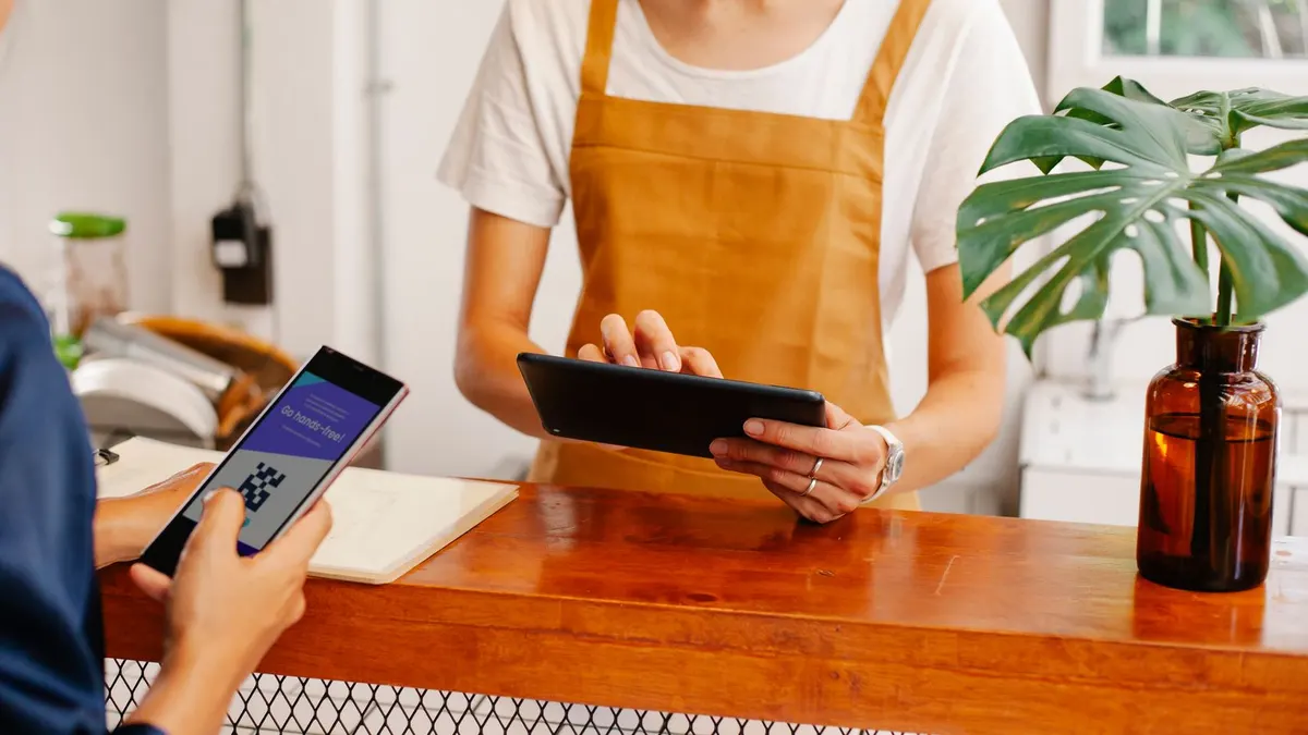 Crop anonymous female employee with application on cellphone screen interacting with partner using tablet at counter in cafeteria